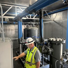 Worker in safety vest and helmet examines control panel in an industrial room with blue pipes and metal tanks.