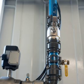 Vertical blue industrial pipeline with metal joints, yellow valve, and attached electronic sensor inside a facility.