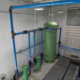 Green industrial tank connected to blue pipes inside a clean facility with white walls and overhead lighting.