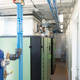 Industrial equipment with blue overhead pipes and green machinery inside a brightly lit, narrow utility room.