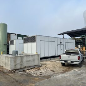A large white industrial unit sits next to a green tank, parked trucks, and factory buildings with smoke in the background.