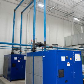 Two large blue industrial machines with vertical blue pipes inside a white-walled facility with a high ceiling.