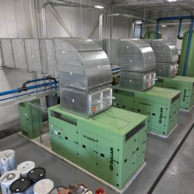 Three large green industrial machines with metal ductwork in a clean, organized facility with barrels and piping nearby.